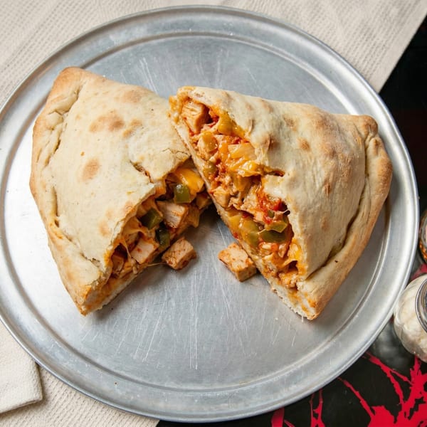 A calzone, cut in half, revealing a filling of chicken, peppers, and cheese on a metal plate.